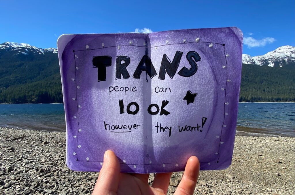 Trans people can look however they want hand written on a purple background in a small sketchbook. Lake and mountains in the background.