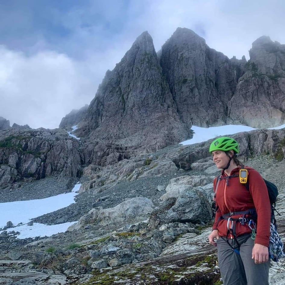 Person with a green helmet, red shirt and grey pants. Tall, jagged mountain in the background. Summer snow in the foreground.