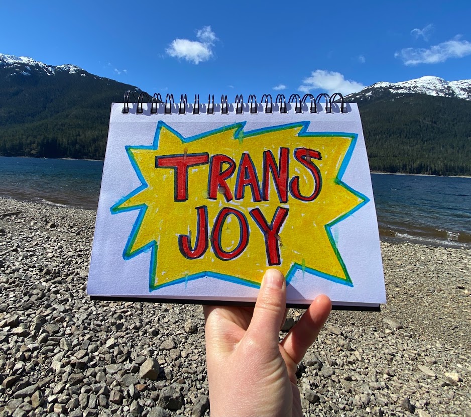 Trans joy drawn in bright markers on white paper. Background is a lake and mountains.